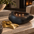 A black stone-shaped candle holder with three lit candles sits on a beige stone surface in a cozy living room setting, alongside a gold decorative object and stacked books.