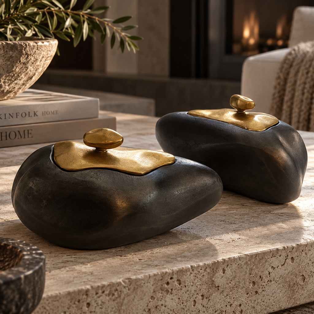 Two black stone-like decorative containers with gold lids sit on a light stone surface in a styled interior setting with books, a plant, and a fireplace in the background.