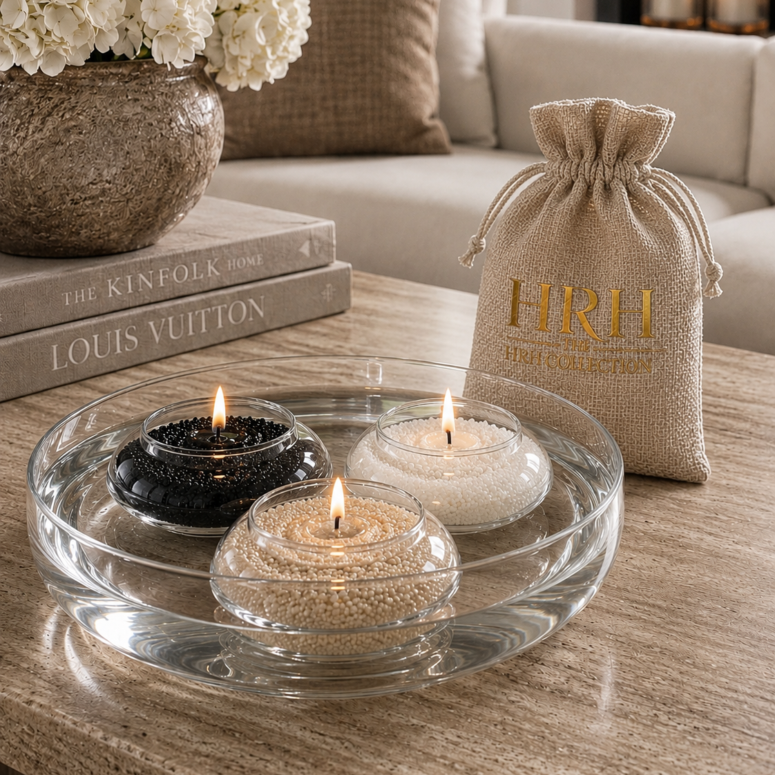 Three lit candle tea light holders in black, white, and clear glass are arranged in a round clear glass tray on a wooden table. A beige burlap drawstring pouch with gold 'HRH First Collection' branding stands beside the tray.