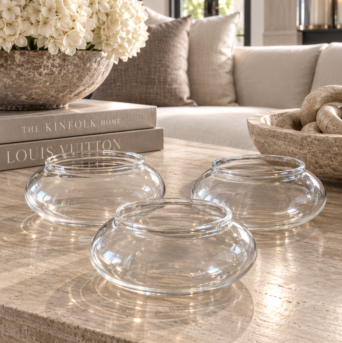 Three clear glass bowl-shaped candle holders arranged on a light stone coffee table in a bright living room setting.