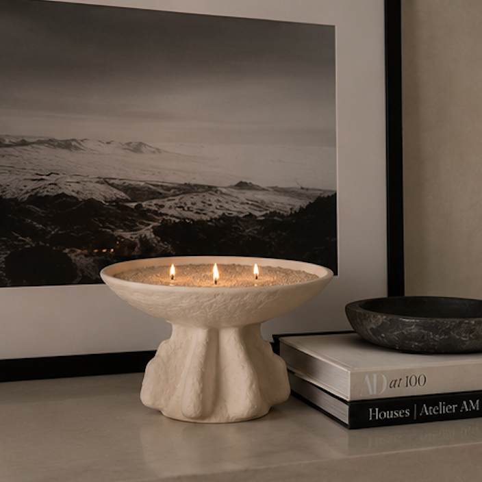 A cream-colored stone pedestal candle holder with three lit candles, styled on a tabletop next to stacked books and a dark stone bowl, with a large black-and-white landscape photograph in the background.
