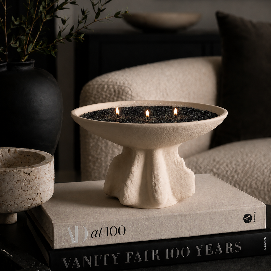 A cream-colored stone pedestal fire pit bowl filled with dark charcoal and three lit candles, styled on top of books in a cozy living room setting.