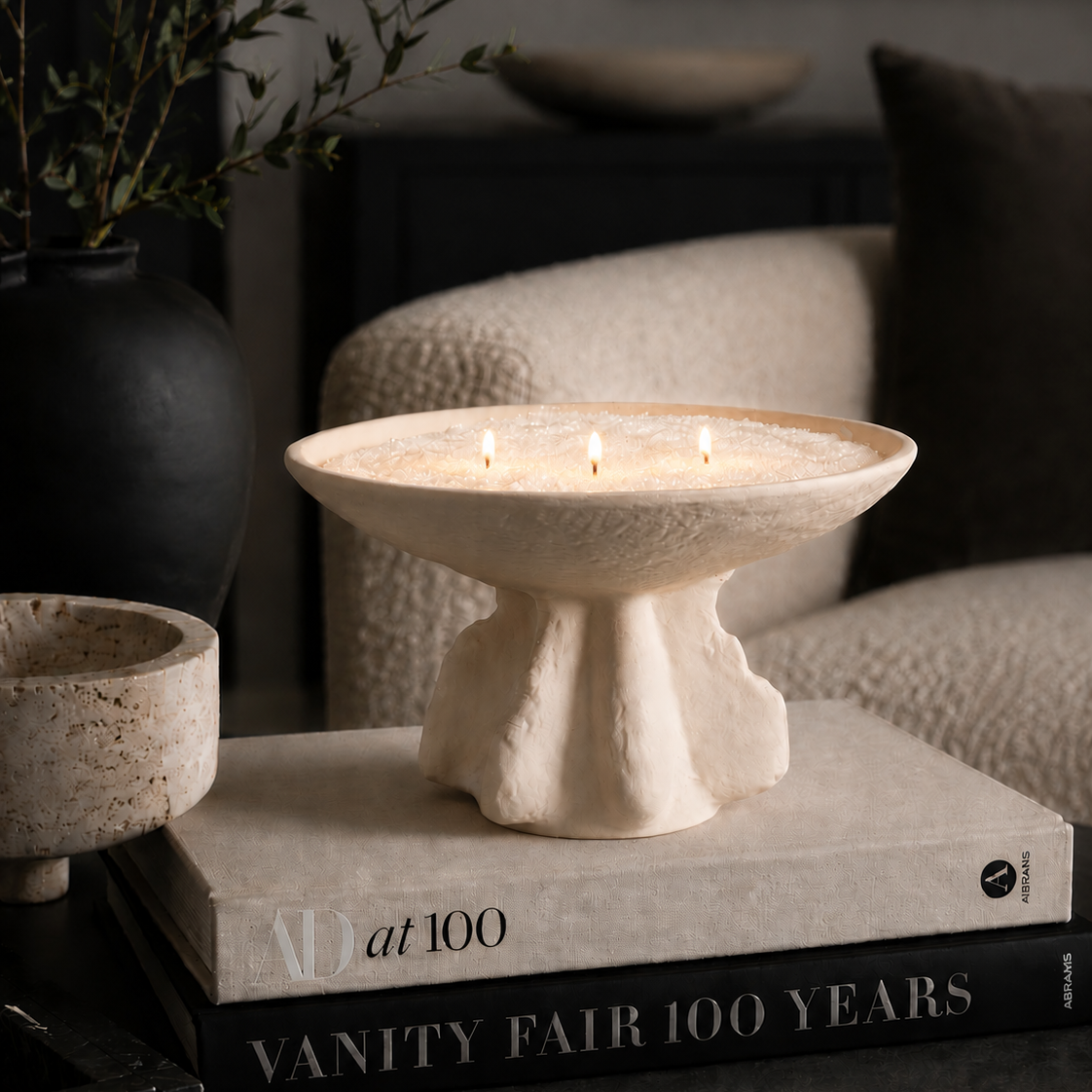 A three-wick ceramic candle holder with a textured, organic sculptural base sits on top of two decorative books in a styled living room setting.