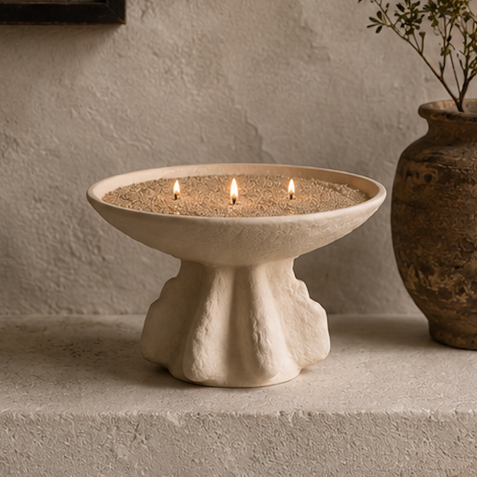 A cream-colored stone pedestal candle holder filled with sand and three lit candles, styled on a textured stone ledge next to a rustic ceramic pot.