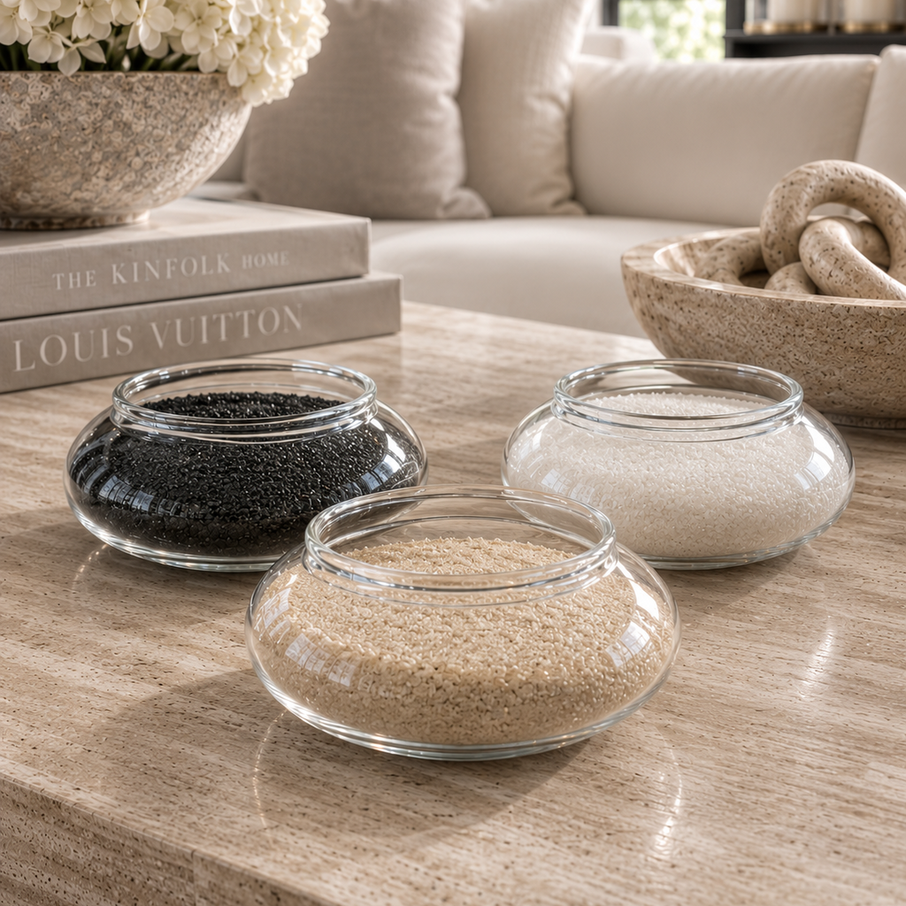 Three round clear glass bowls filled with black, beige, and white decorative stones or beads, arranged on a light wood table in a styled living room setting.