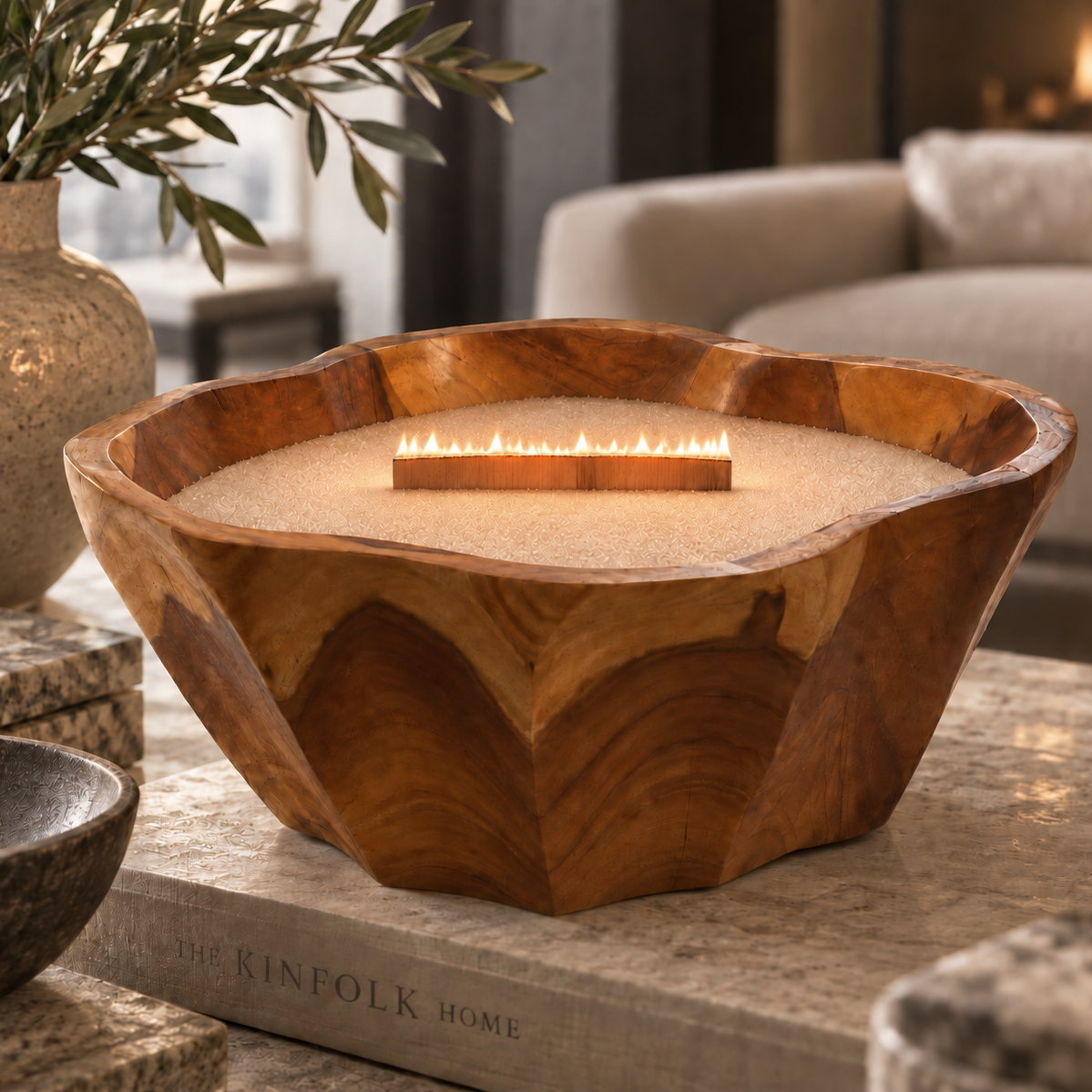 A large carved wooden fire pit bowl filled with sand, with a small flame visible in the center, styled on a stone surface in a cozy living room setting.