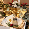 A holiday gift box filled with HRH brand products, including three gold pouches, a small burlap bag with a gold seal, and decorative holiday elements like pine branches, red berries, and white beads, set on a countertop with a blurred festive background.