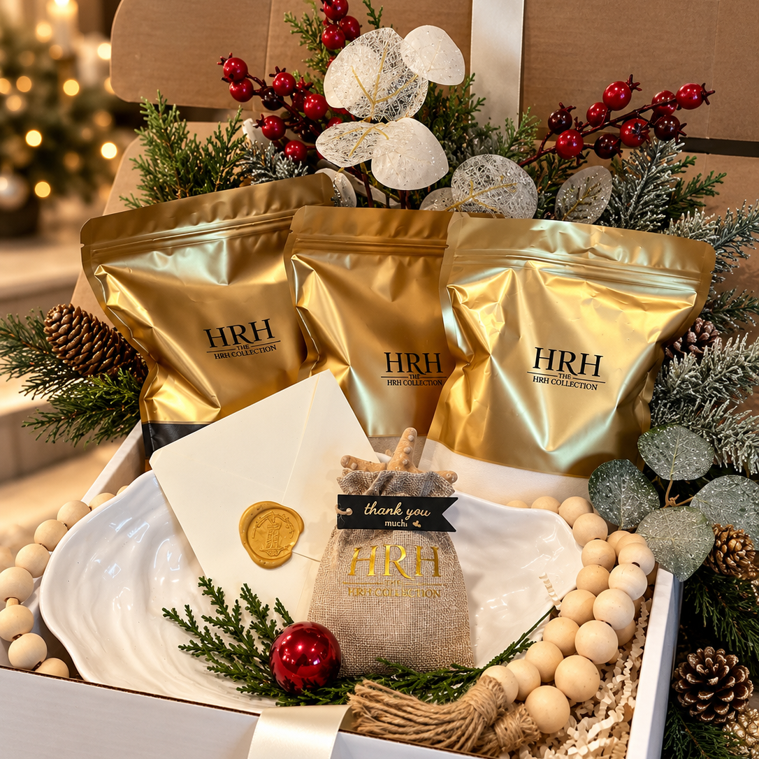 A holiday gift box filled with HRH brand products, including three gold pouches, a small burlap bag with a gold seal, and decorative holiday elements like pine branches, red berries, and white beads, set on a countertop with a blurred festive background.