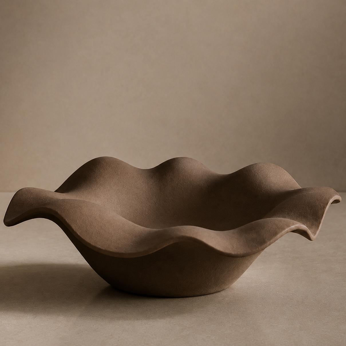A wavy, scalloped ceramic bowl in a matte taupe-brown glaze, set against a matching warm beige background.
