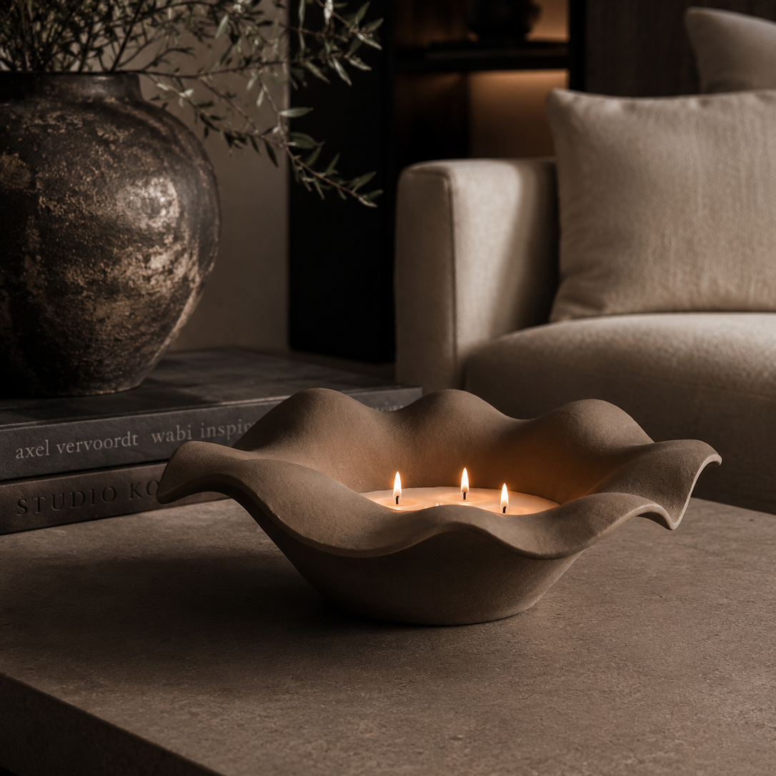 Wavy ceramic candle holder with three candles on a dark surface, with books and decorative items in the background.
