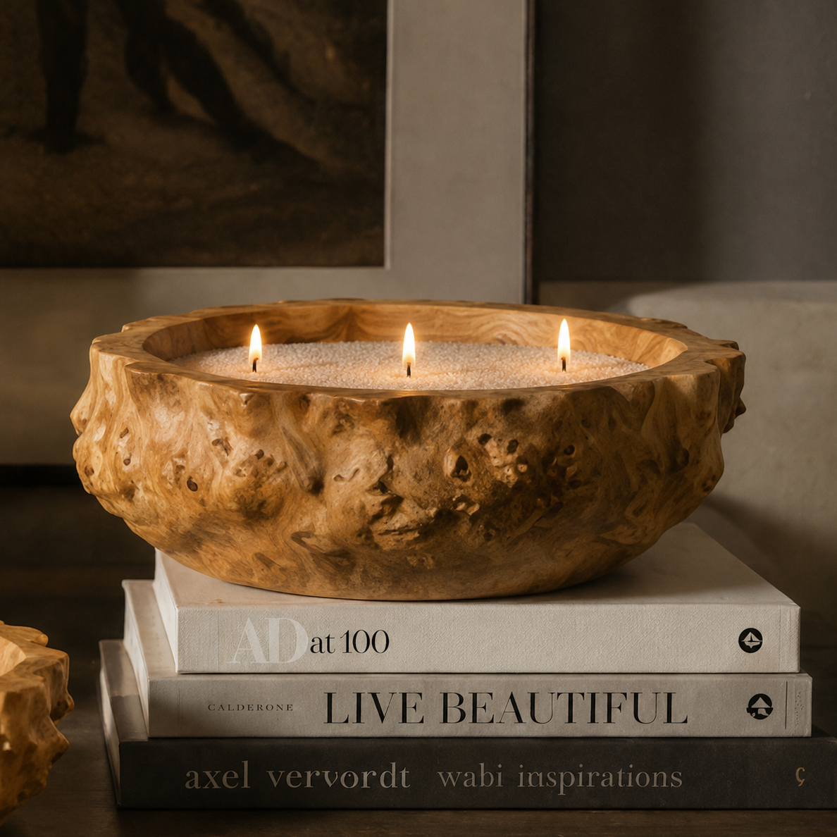 A rustic carved wooden bowl candle holder with three lit candles, styled on a stack of books in a warm, moody interior setting.