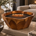 A wooden fire pit bowl with a carved, organic shape, filled with black gravel and a small flame, styled on a stone table in a cozy living room setting.