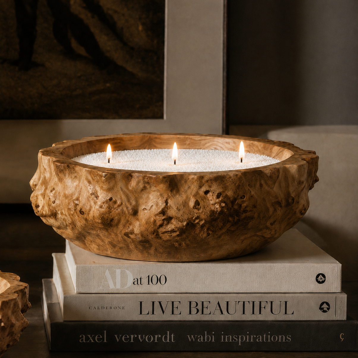A three-wick soy wax candle in a rustic, organic-shaped wooden bowl, styled on top of three books in a warm, dimly lit interior setting.