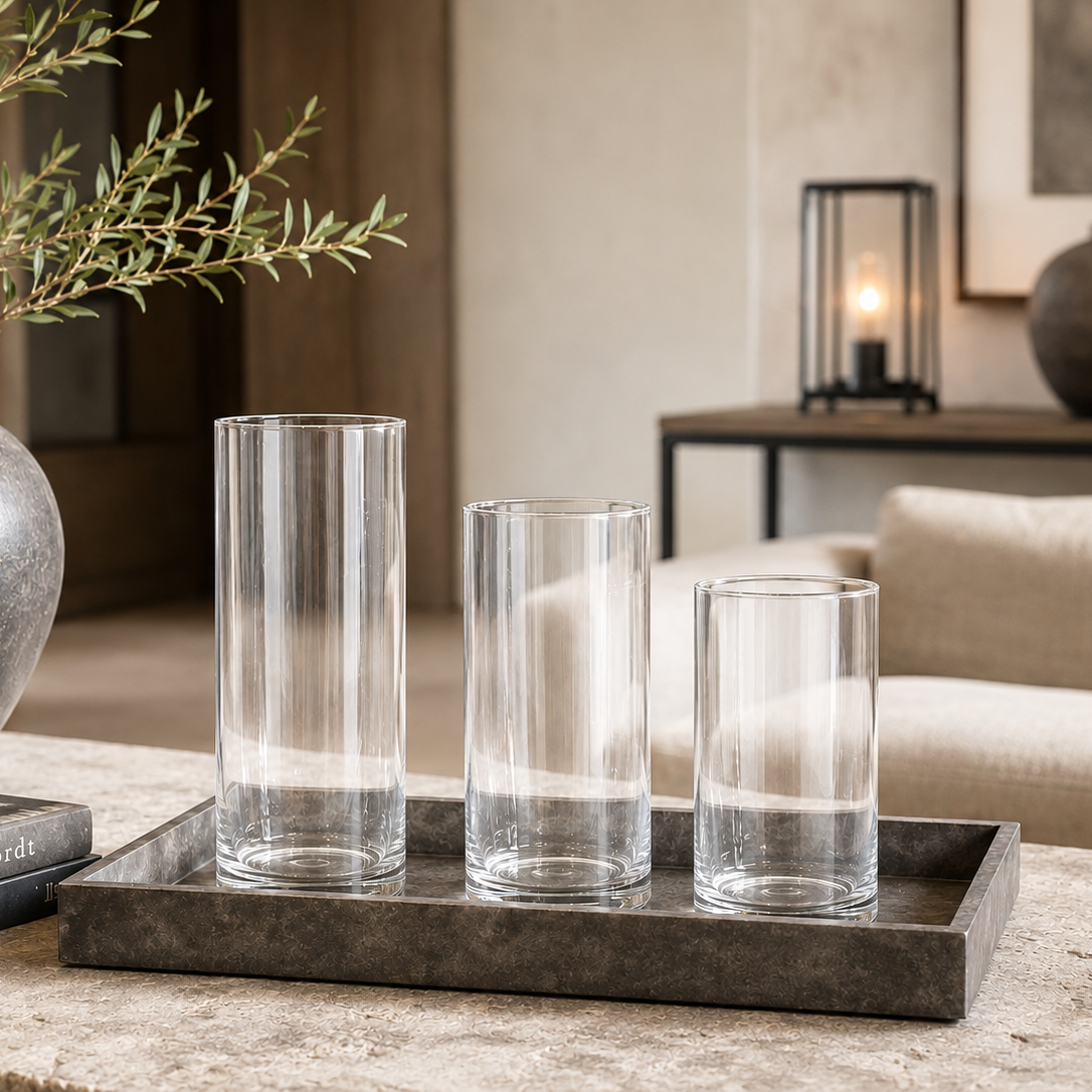 Three clear glass cylinder vases of different heights displayed on a dark rectangular tray in a styled living room setting.