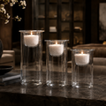 Three clear glass cylindrical candle holders with white candles inside, arranged on a dark stone surface in a dimly lit interior setting.