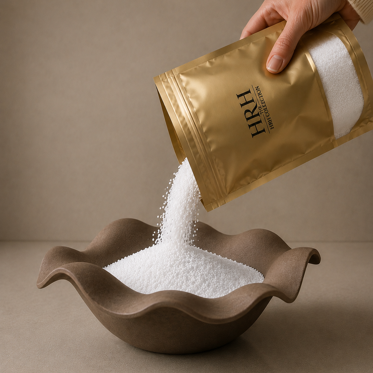 A hand pours white granulated sugar from a metallic gold pouch labeled 'HRH' into a brown ceramic bowl against a neutral beige background.