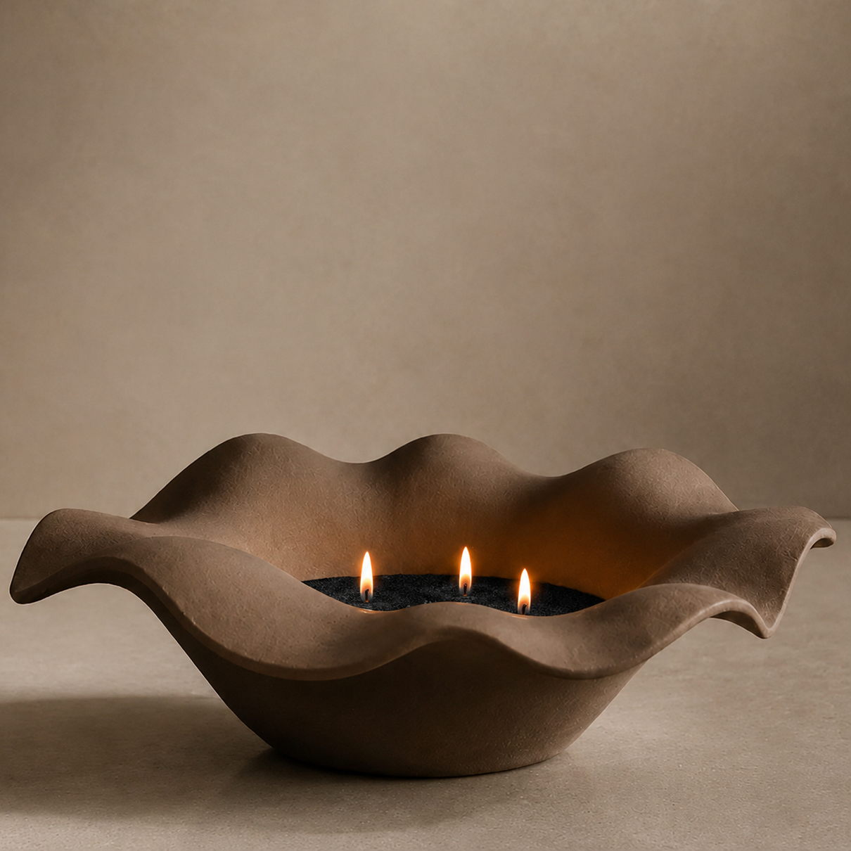 A wavy, flower-shaped ceramic candle holder in a warm taupe-brown color, containing three lit candles on a matching textured surface against a neutral beige background.