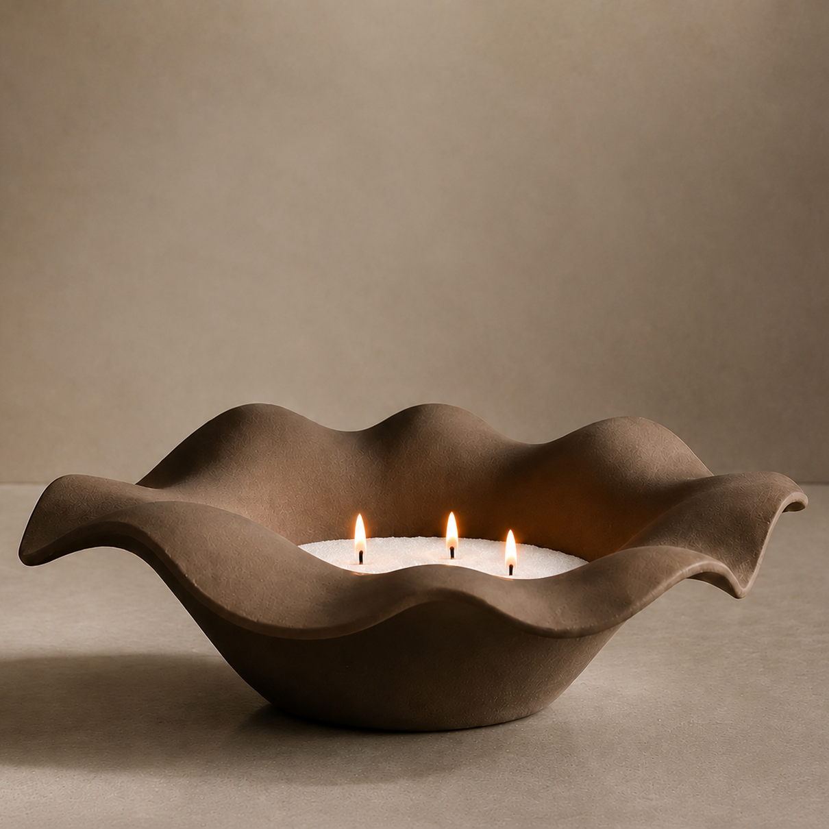 A sculptural, wavy ceramic candle holder in a warm taupe-brown color, holding three white tea light candles with lit flames, set against a matching neutral studio background.